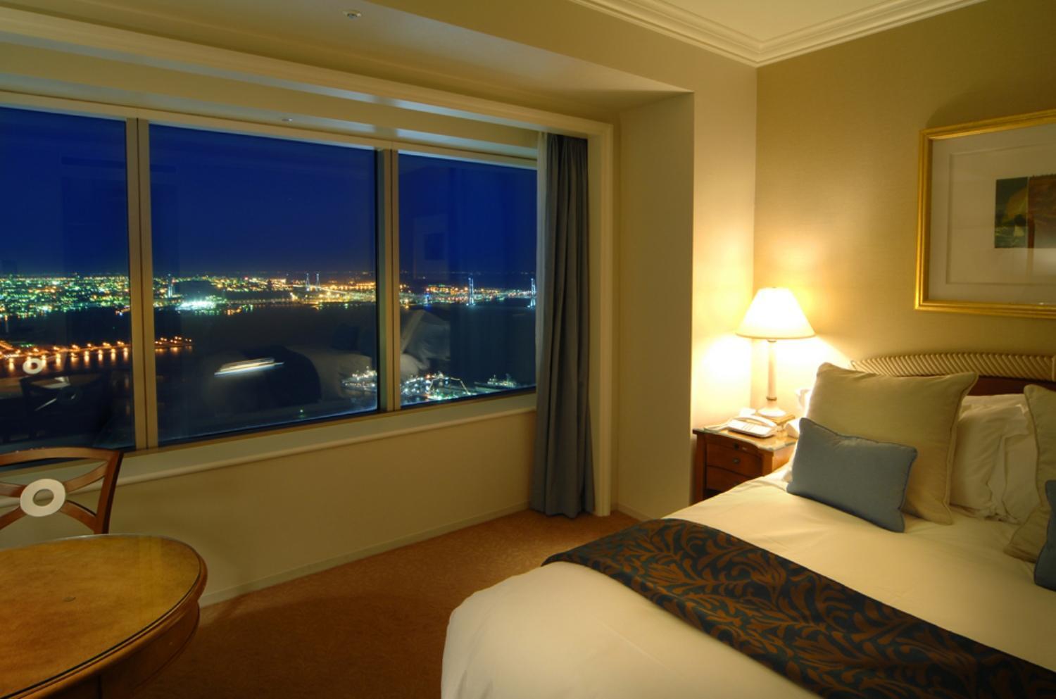 Yokohama Royal Park Hotel Room photo The photo shows a hotel room at night with a large window displaying a view of a city illuminated by lights. The room features a neatly made bed with decorative pillows and a dark throw blanket. There is a bedside table with a lamp providing warm lig