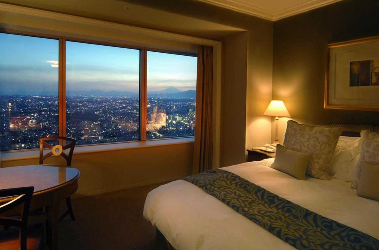 Yokohama Royal Park Hotel Room photo The photo shows a hotel room with an elegant and cozy atmosphere. There is a large bed with decorative pillows and a comforter, positioned next to a big window that offers a stunning view of a cityscape at dusk. The room features a table and chair, l