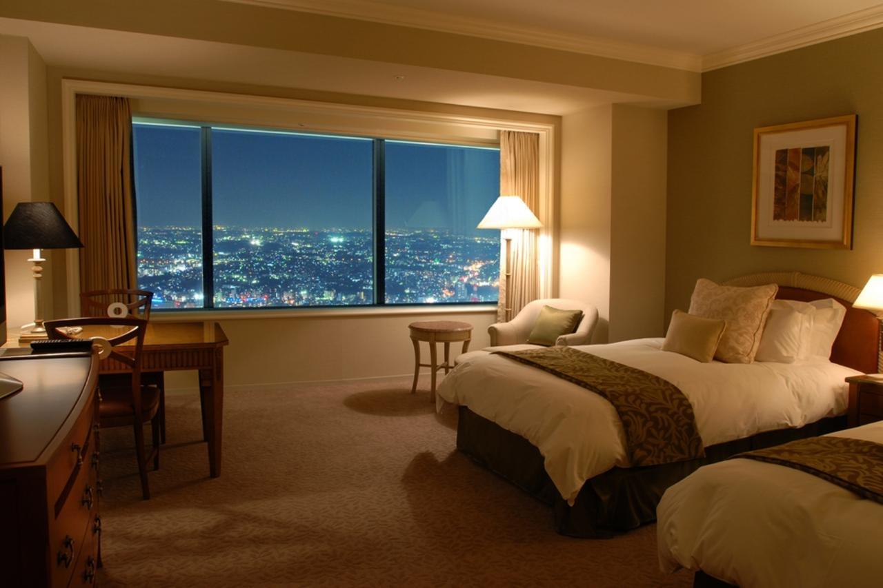 Yokohama Royal Park Hotel Room photo The photo shows a hotel room with two beds, neatly made with white linens and decorative pillows. There is a chair and a small table next to one of the beds. The room features a large window that offers a view of a cityscape lit up at night, with lig